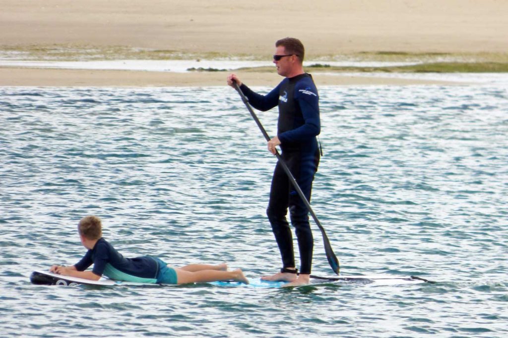 A Man paddle boarding with his son lying down on the board as well